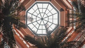 Blick auf eine geometrische Glasdecke mit einem zentralen achteckigen Design, umgeben von einer symmetrischen Struktur aus Holzträgern und Palmenwedeln.