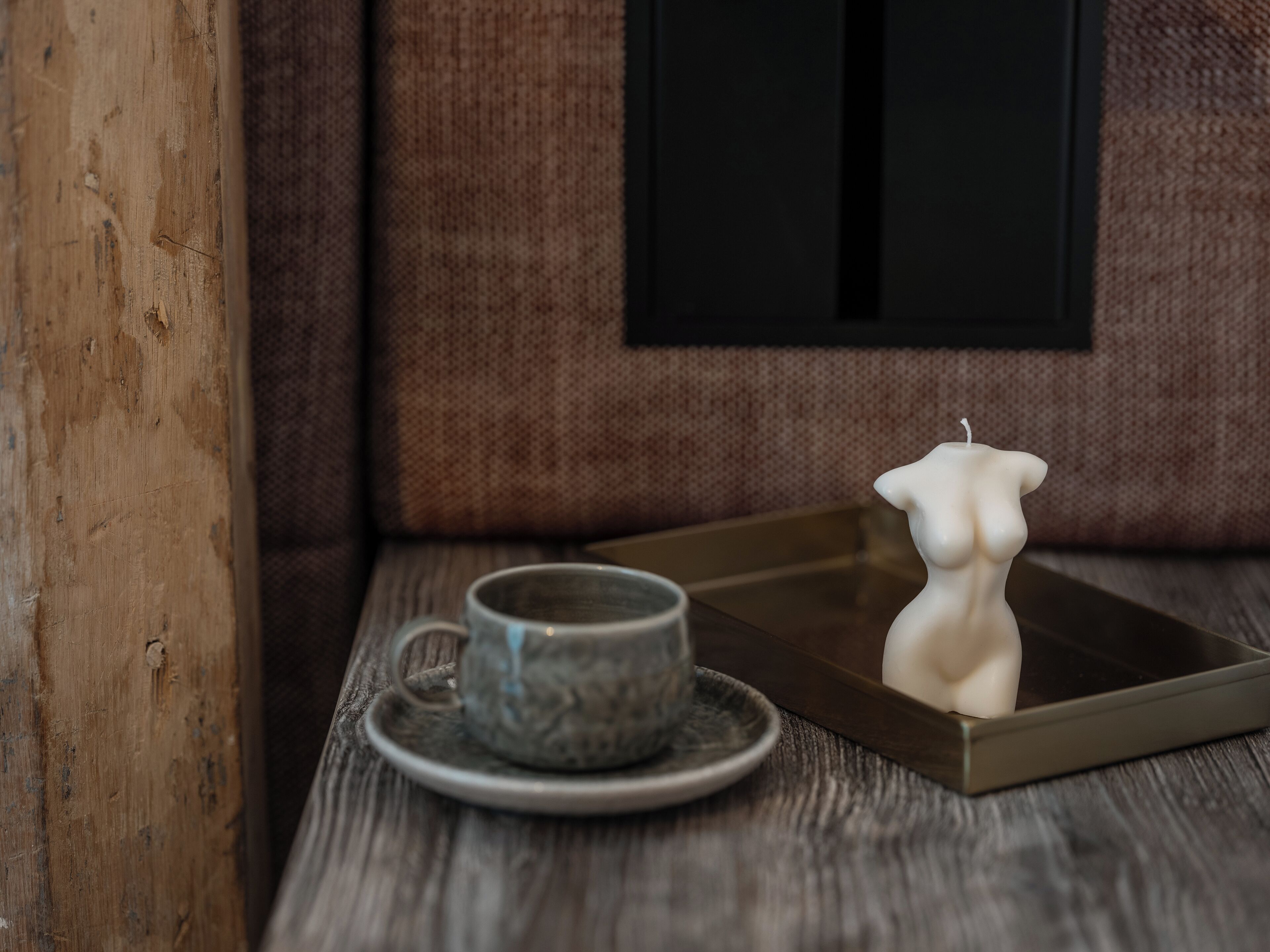 A wooden table with a ceramic cup and saucer, a white candle, and a framed picture on the wall.