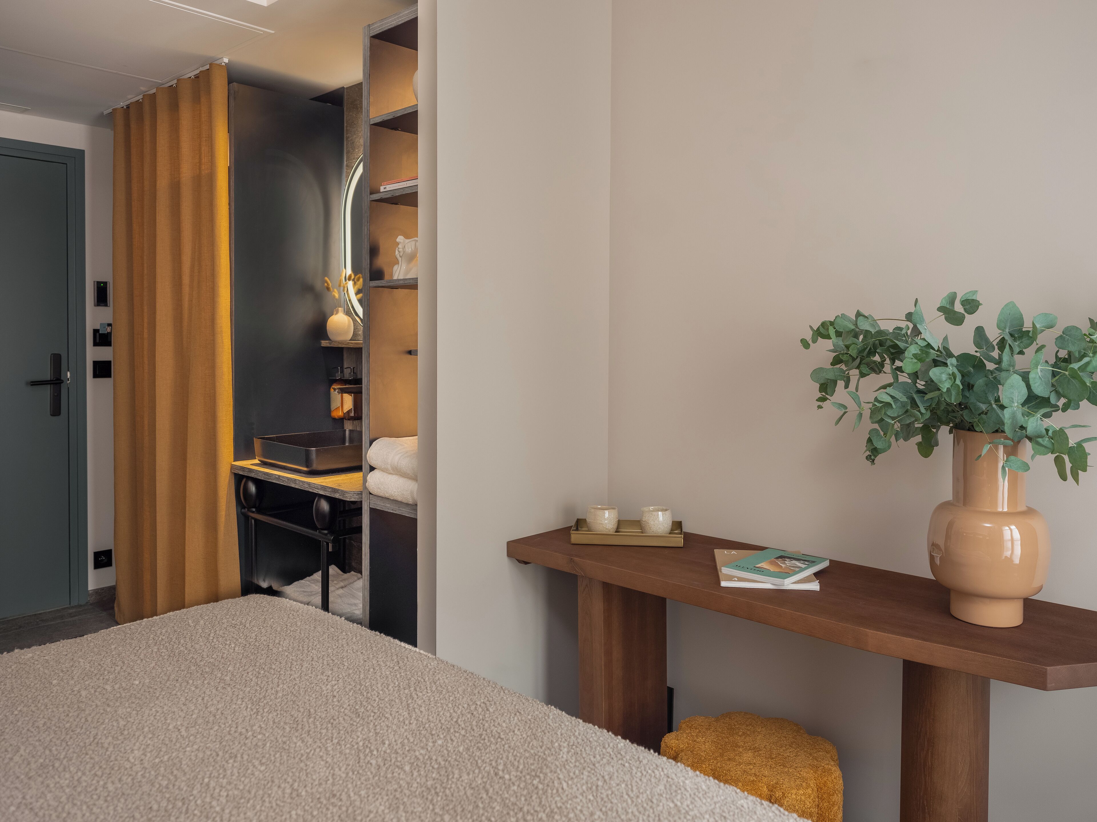 A modern hotel room with a bed, a wooden nightstand, a beige carpet, a brown curtain, a bookshelf, and a potted plant.