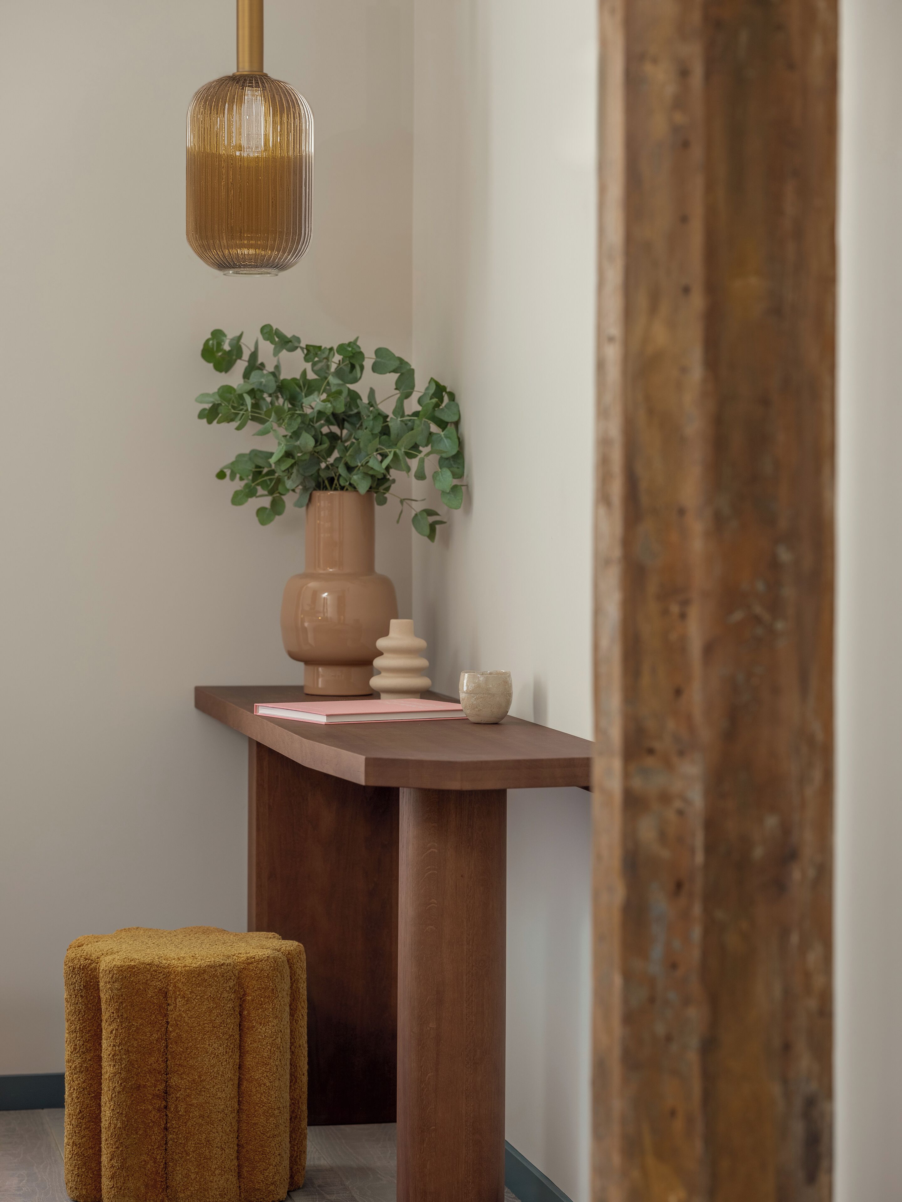 A corner of a room with a wooden table, a potted plant, a small stool, and a hanging light fixture.