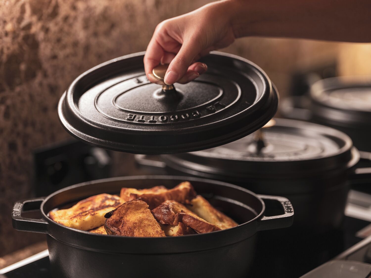 Une main soulève le couvercle d’une cocotte en fonte noire posée sur une cuisinière, et on peut voir de la nourriture à l’intérieur.