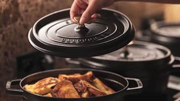 Une main soulève le couvercle d’une cocotte en fonte noire posée sur une cuisinière, et on peut voir de la nourriture à l’intérieur.