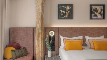 A hotel room with a large bed, a seating area with cushions, and two framed artworks on the wall.