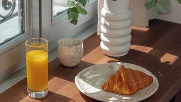 Morgenmadsbord med et glas appelsinjuice, en croissant og en hvid keramikkop på et træbord ved et vindue.