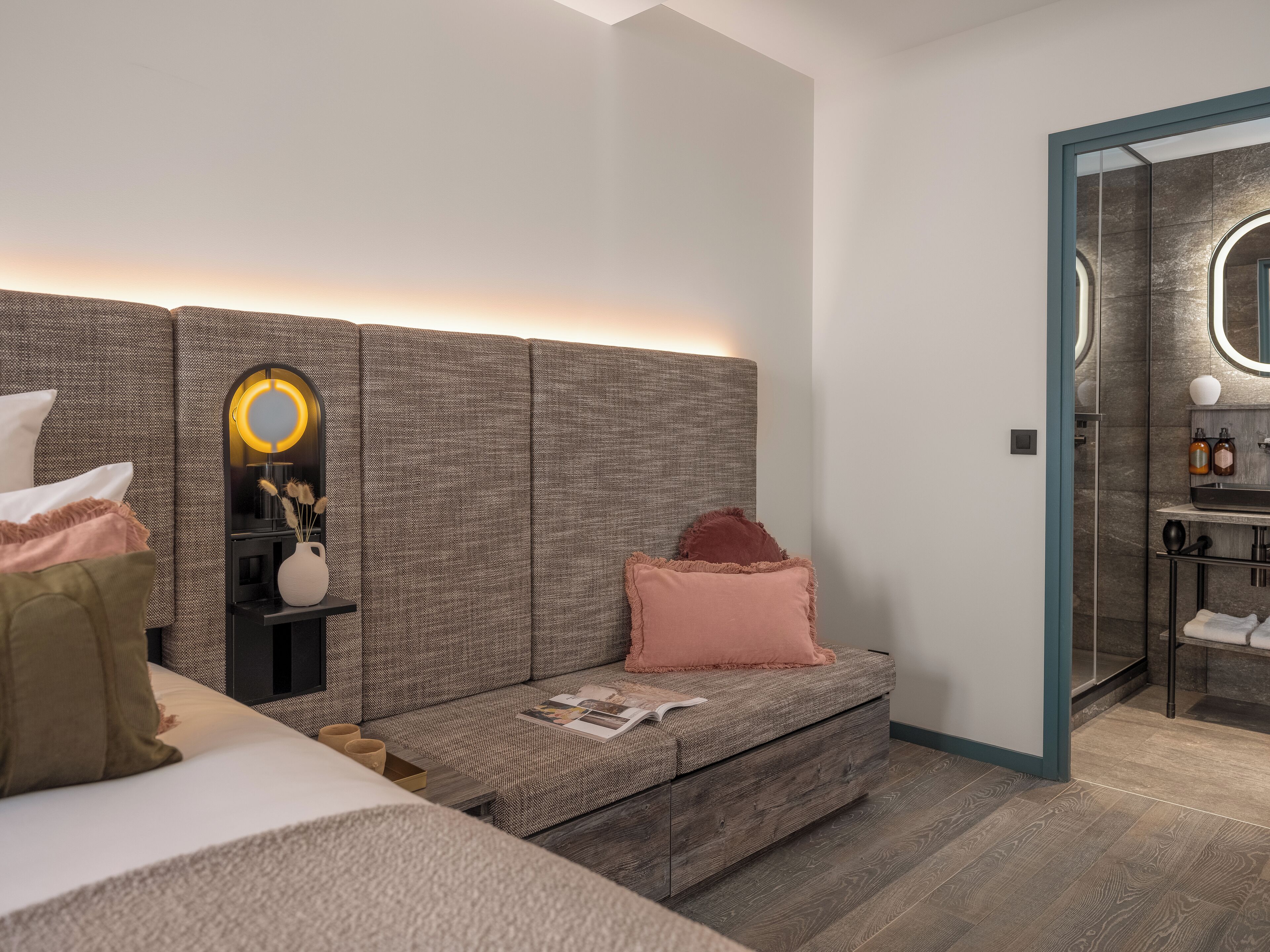 A modern bedroom with a large bed, a bedside table with a mirror, a small seating area, and a view into the bathroom.
