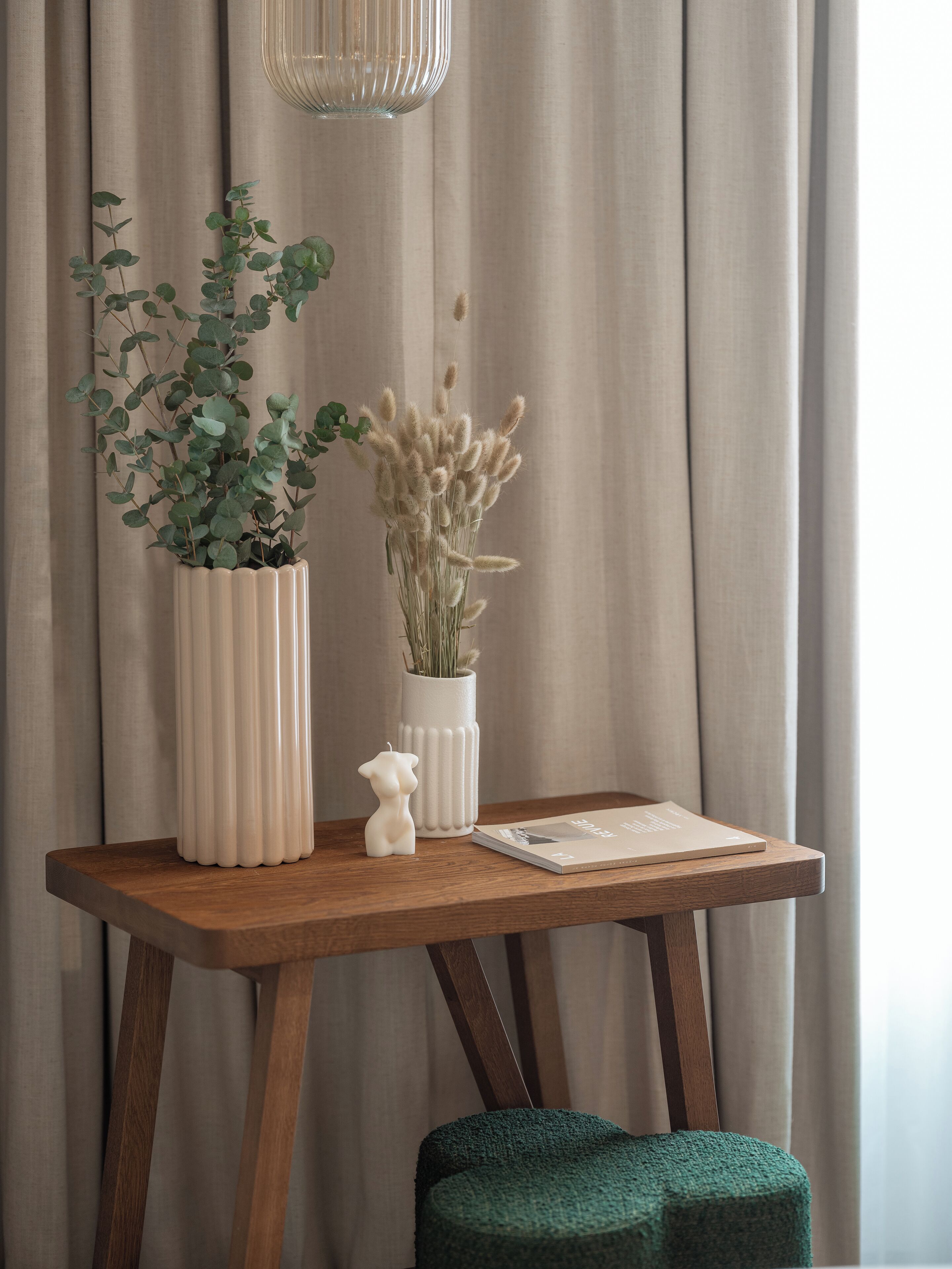 A wooden table with two white vases, one with green foliage and the other with dried flowers, and a small white figurine.