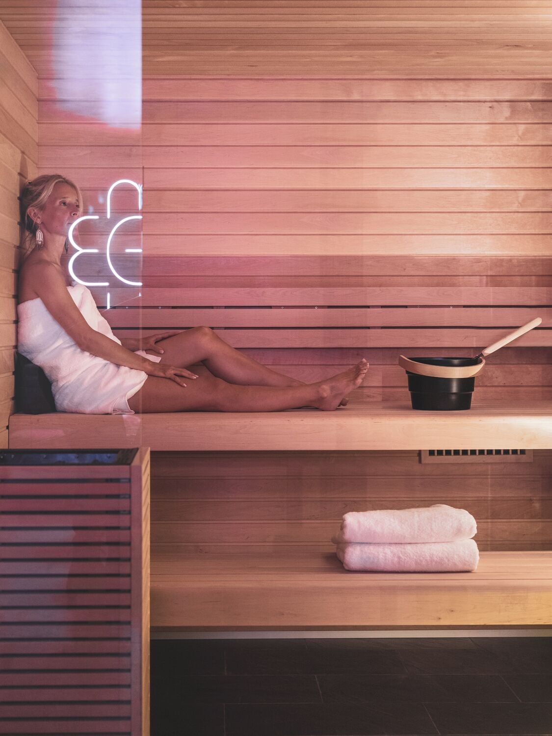Una persona sentada en una sauna de madera, con una toalla y una olla sobre una repisa.