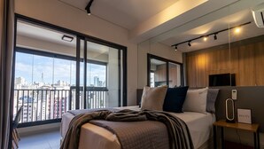 A modern hotel room with a large bed, a bedside table with a lamp, a TV, and a city view through large windows. - Vossa Bossa Facto (São Paulo)