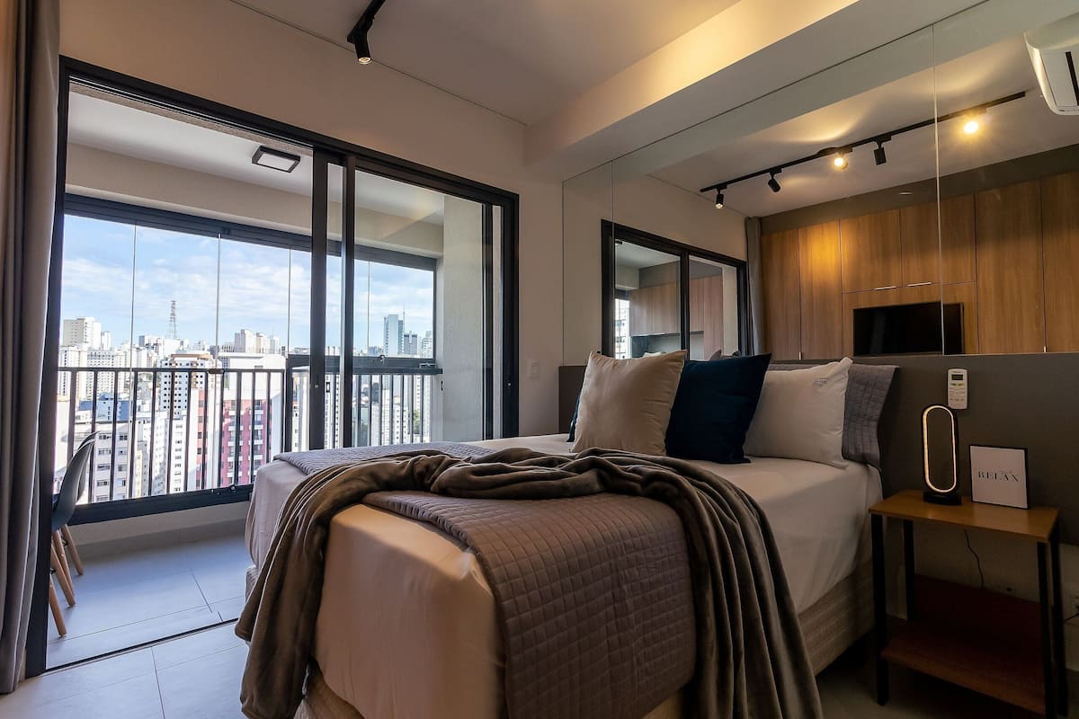 A modern hotel room with a large bed, a bedside table with a lamp, a TV, and a city view through large windows.
