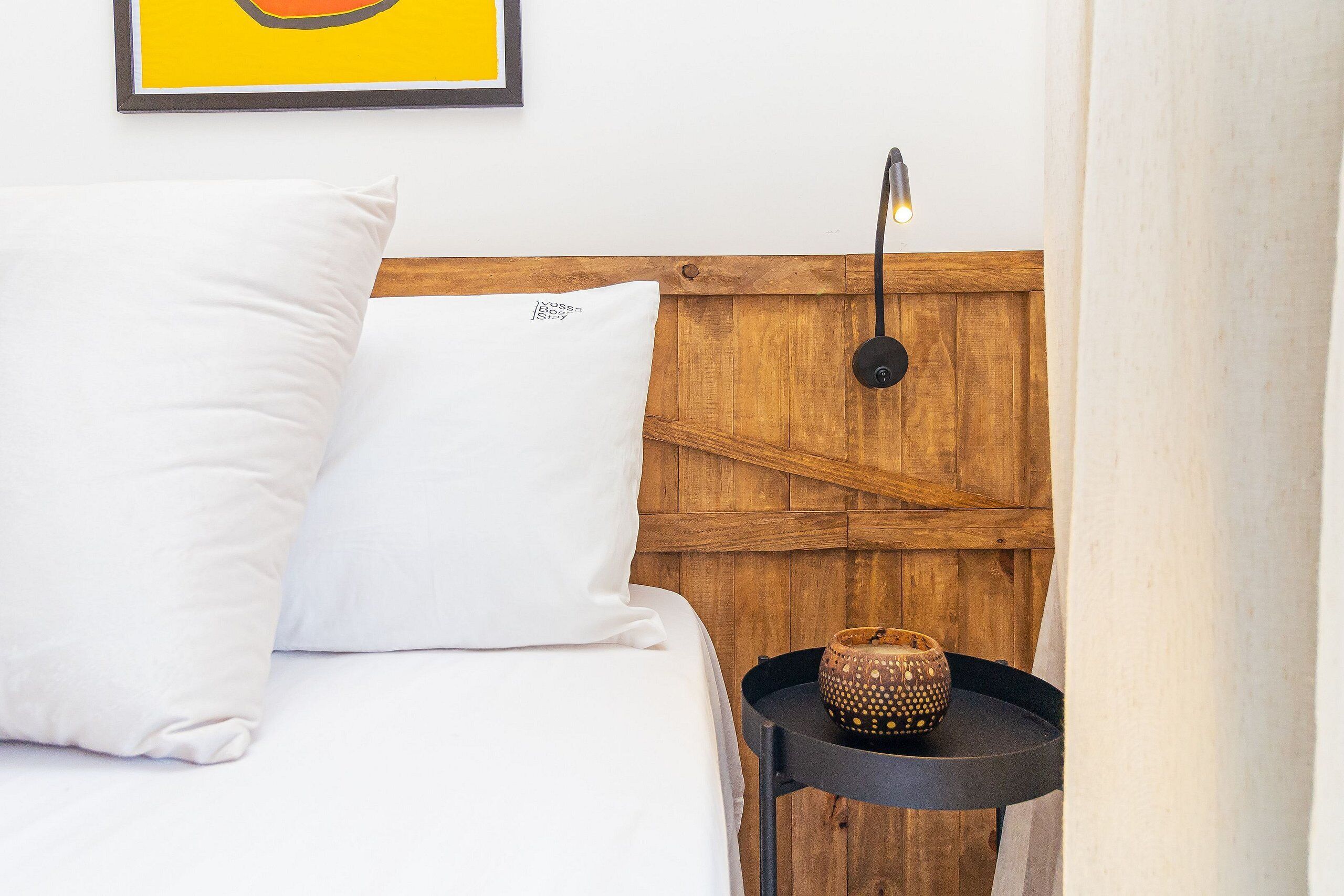 Una cama bien hecha con ropa de cama y almohadas blancas, un cabecero de madera, una mesita de noche negra pequeña con un objeto decorativo y un cuadro en la pared.