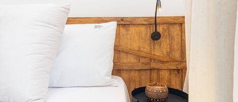 Una cama bien hecha con ropa de cama y almohadas blancas, un cabecero de madera, una mesita de noche negra pequeña con un objeto decorativo y un cuadro en la pared.