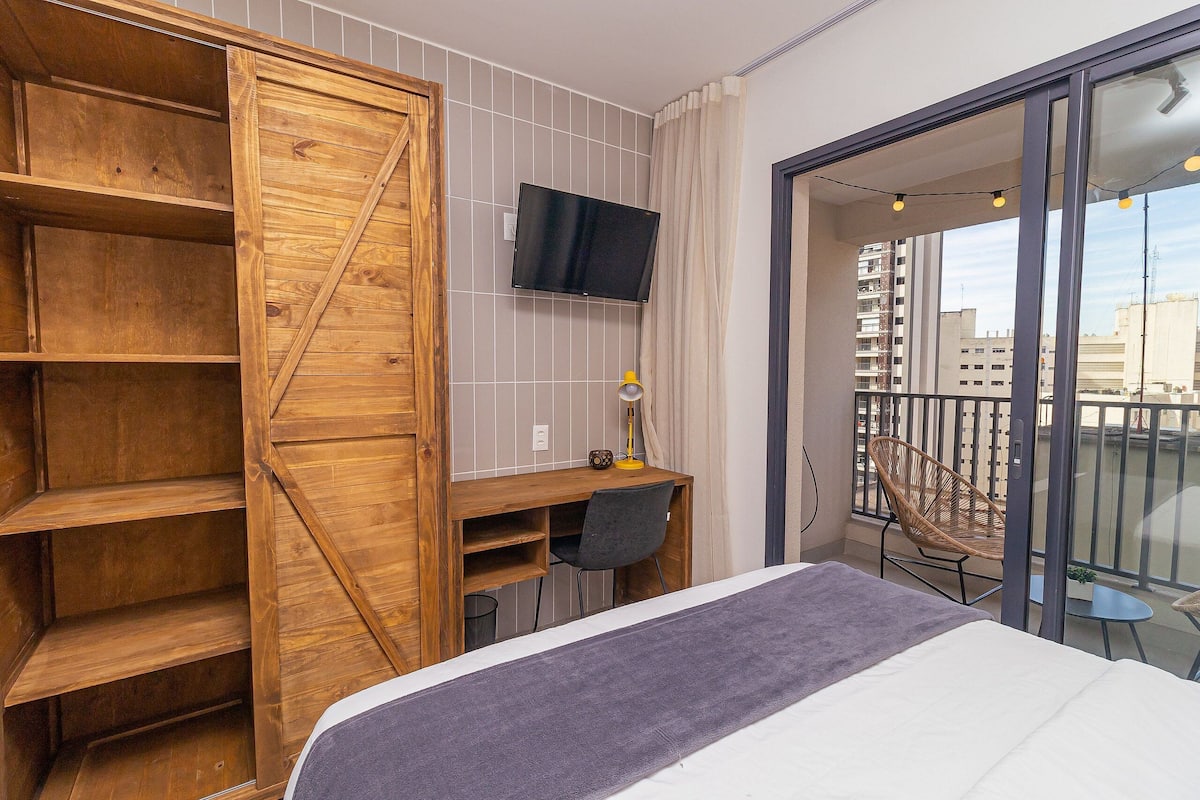 A bedroom with a bed, a wooden wardrobe, a desk with a chair, a TV, and a balcony with a chair and table.