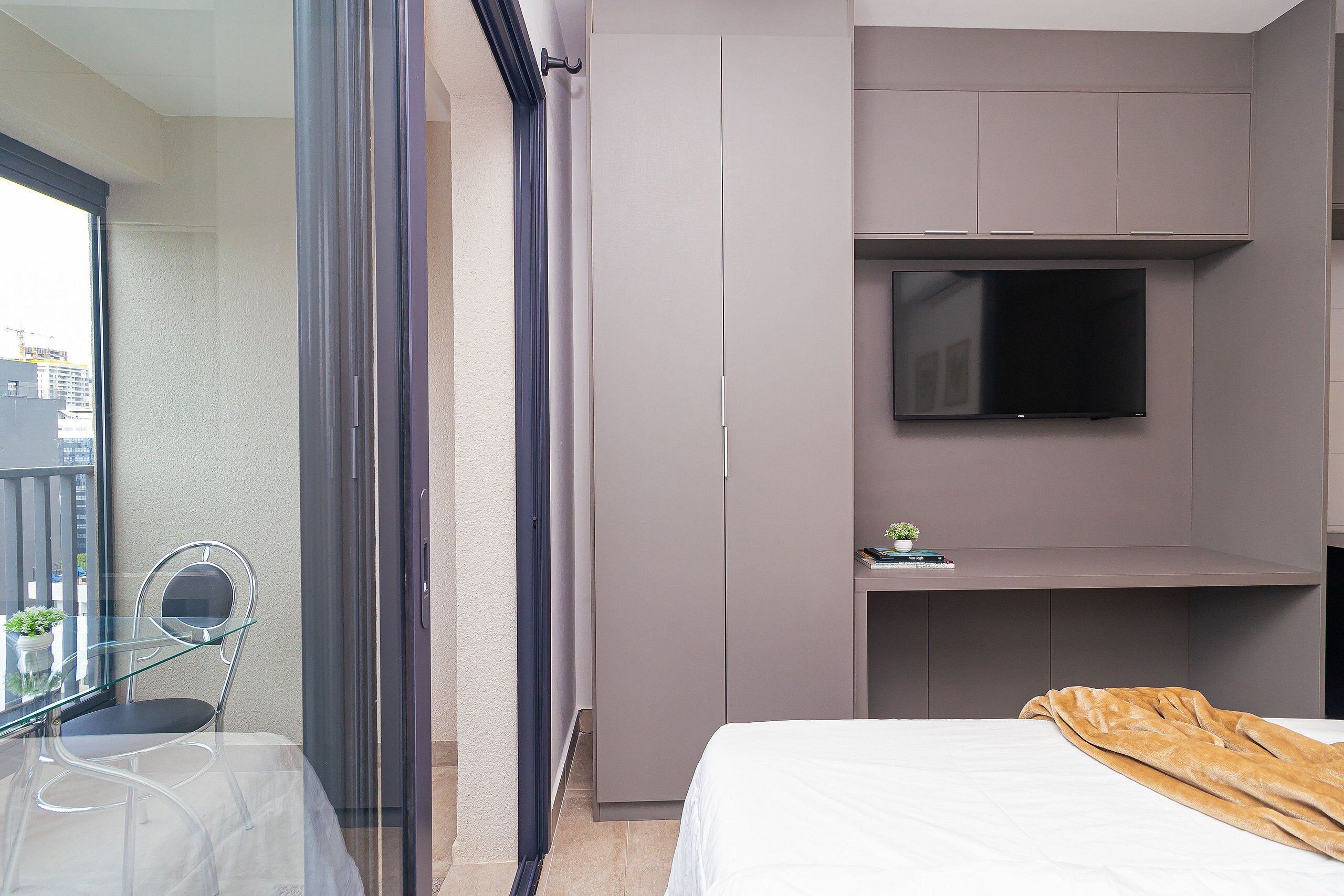 Moderni hotellihuone, jossa on sänky, seinälle asennettu televisio, lasinen pöytä ja tuoli sekä liukuovi.