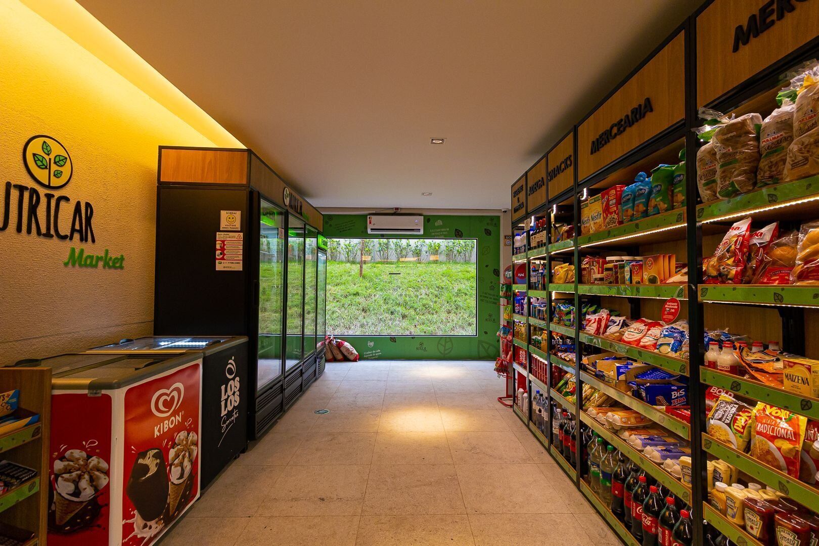 Interno di un negozio di alimentari con scaffali pieni di diversi prodotti, una vetrina refrigerata e vista su una zona erbosa attraverso una parete di vetro.