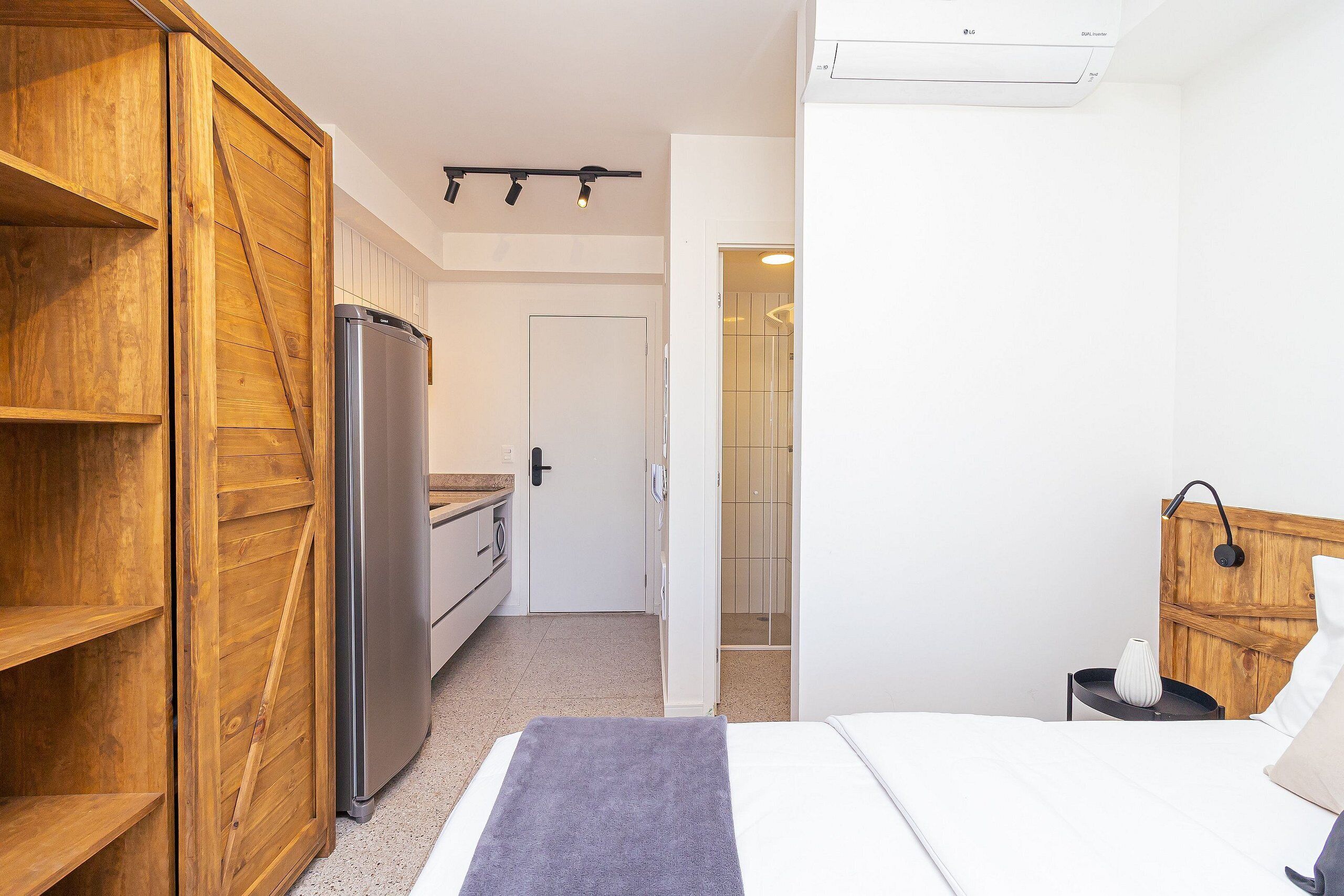 A bedroom with a bed, a wooden wardrobe, a small bedside table, and a bathroom visible through an open door.