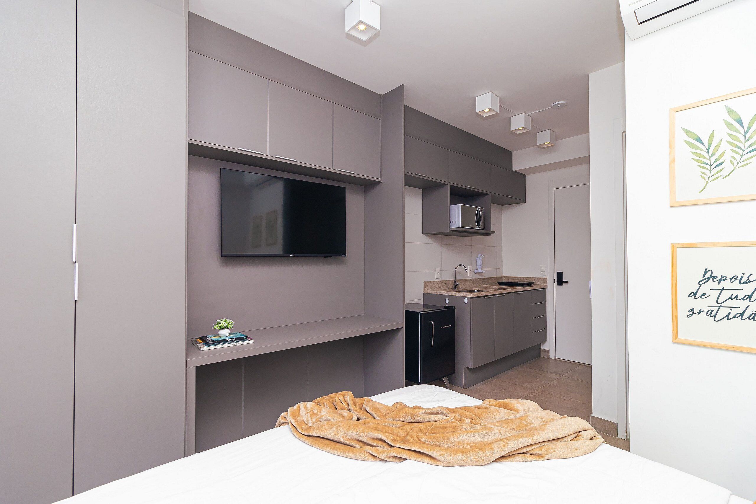A modern bedroom with a bed, a bedside table, a TV mounted on the wall, and a small kitchenette area.