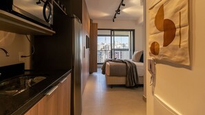 A modern kitchen with a built-in oven, a dining area with a table and chairs, a large window with a city view, and a bedroom with a bed and a bedside table. - Vossa Bossa Facto (São Paulo)
