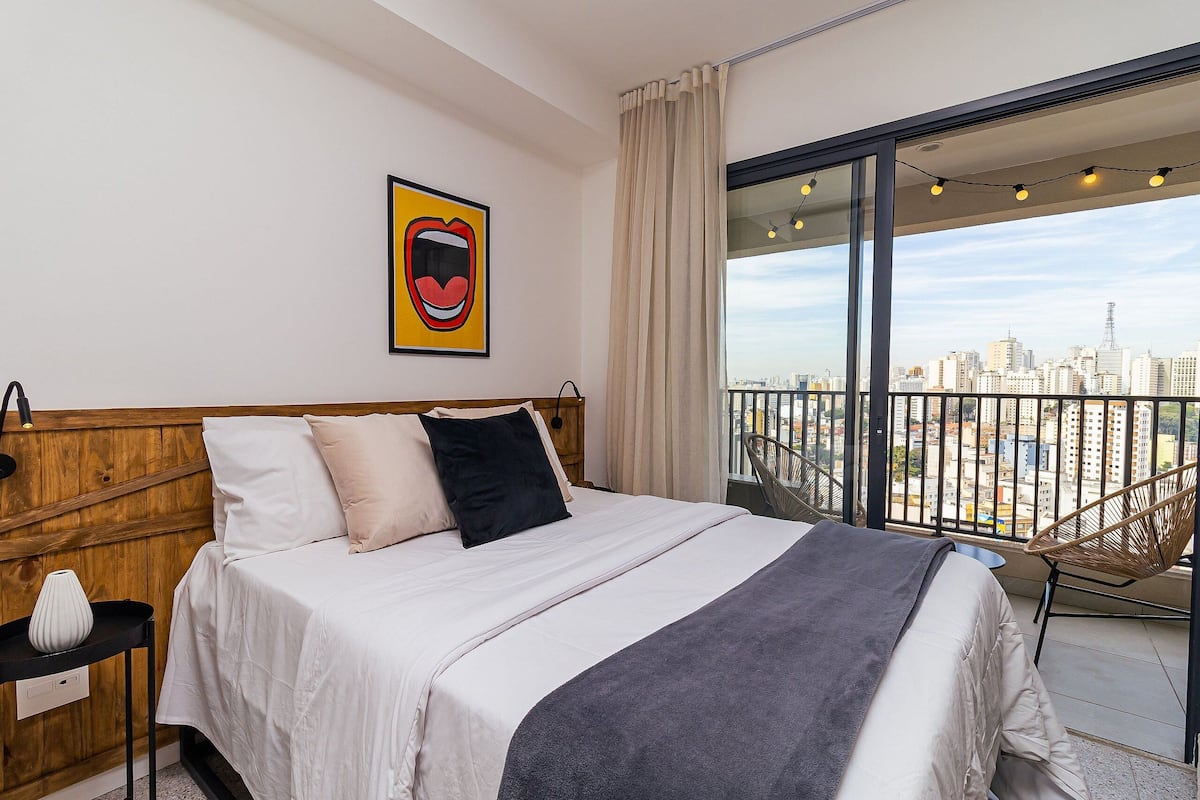 A bedroom with a bed, a nightstand, a chair, a city view balcony, and a framed artwork on the wall.