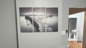 Una habitación con un sofá negro, una obra de arte de tres paneles en la pared que representa un muelle, y suelo de madera.