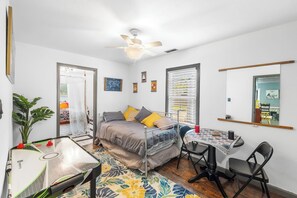 Une chambre à coucher de taille modeste, comprenant un lit, un bureau, une chaise, un ventilateur de plafond et une fenêtre avec des stores.