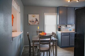 Une cuisine de taille réduite avec une table à manger, des chaises, un panier de fruits et un tableau accroché au mur.