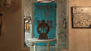 Un couloir avec une porte turquoise, une table ronde ornée d’une sculpture et une lampe fixée au mur.