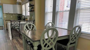 Un comedor con una mesa de madera y cuatro sillas, una cocina con armarios blancos y un salpicadero de azulejos, y un ventanal con persianas.
