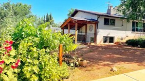 A house with a covered porch, a garden with red flowers, and a clear blue sky. - Central & Peaceful Getaway at Cozy Studio Apt.  (Toquerville)