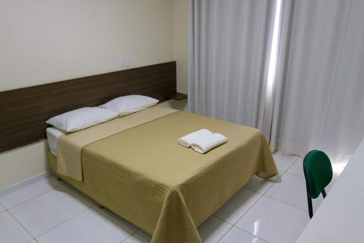 A neatly made bed with a beige bedspread and white pillows, a wooden headboard, and a green chair in a clean, white room with a curtain on the window.