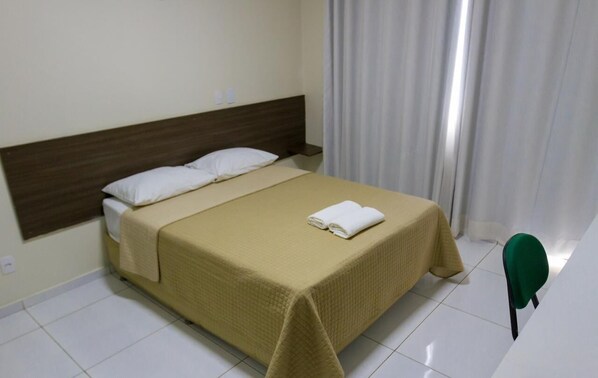 Una cama bien hecha con colcha color beige y almohadas blancas, cabecero de madera y una silla verde en una habitación limpia y blanca con cortina en la ventana.