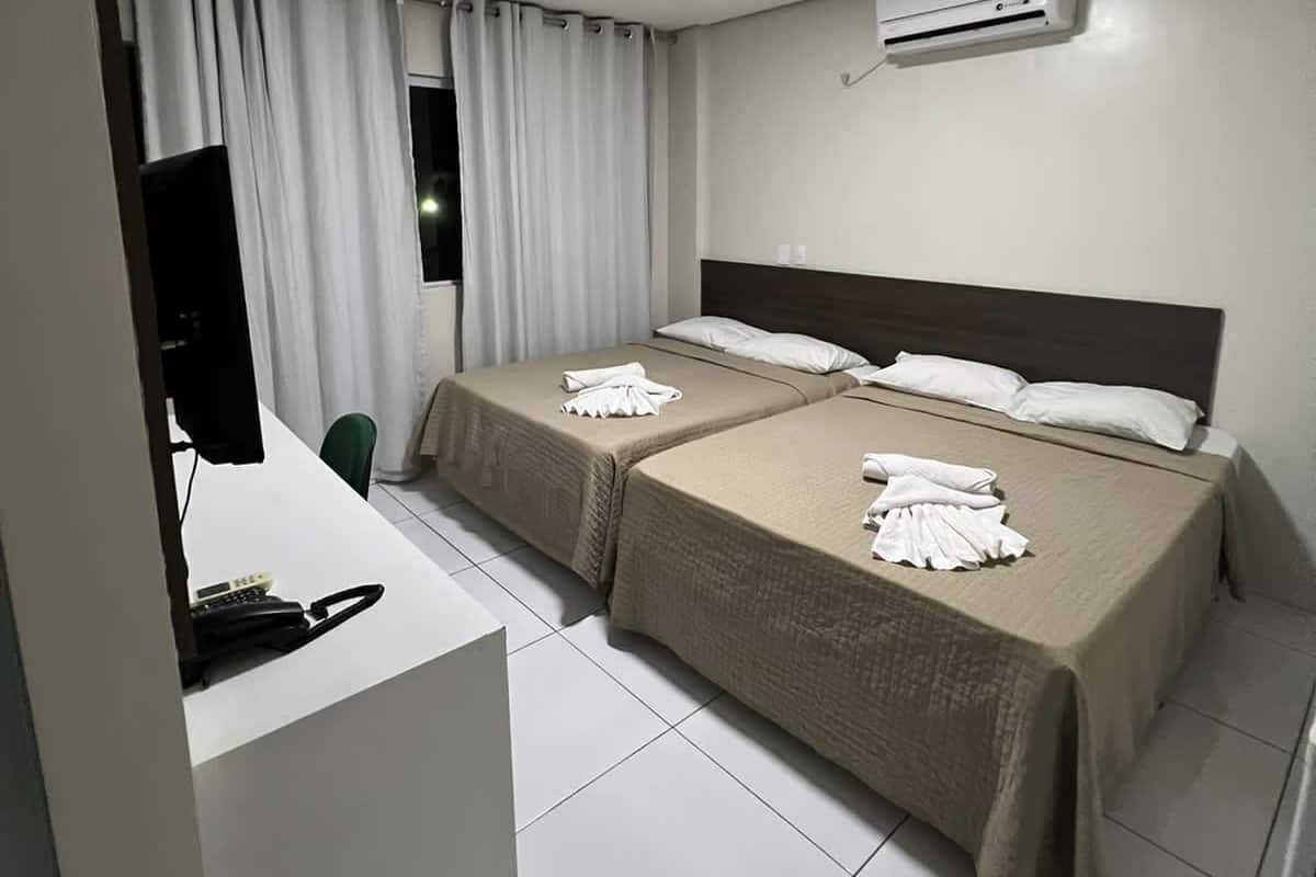 A hotel room with two beds, a desk with a computer and phone, a TV, and an air conditioning unit.