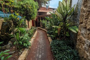 Ein schmaler Ziegelweg, gesäumt von üppigen tropischen Pflanzen und einer Steinmauer, führt zu einem Gebäude mit einem Balkon.
