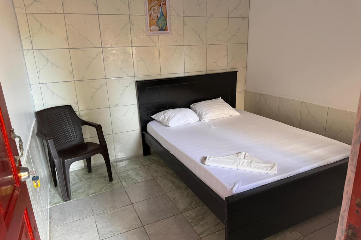 A neatly made bed with white linens, a black headboard, and a single white pillow. A plastic chair is placed against the wall, and a small framed picture hangs above the bed.