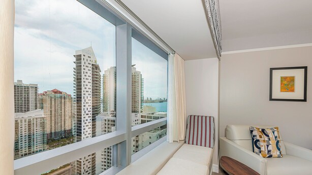 A modern hotel room with a large window offering a city view, a white sofa with a patterned pillow, and a small wooden table. - Elegant King Studio Suite + Kitchenette (Miami)