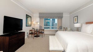 A hotel room with a large bed, a television, a desk with chairs, a window with a view of buildings, and a bench. - Elegant King Studio Suite + Kitchenette (Miami)