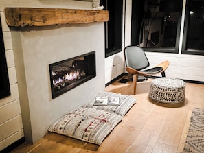 A cozy room with a fireplace, a chair, a patterned ottoman, and a rug.