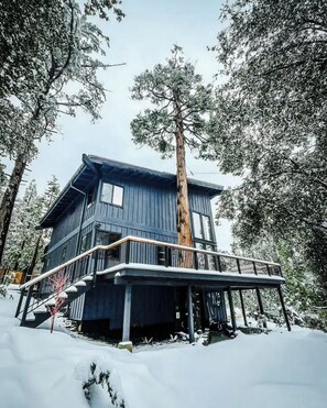 A two-story cabin with a balcony, surrounded by snow and trees. - Cedar Haus Modern Family Haven w Game Room, Hot Tub, EV Charger, Fully Fenced Yard, Dogs OK (Idyllwild-Pine Cove)