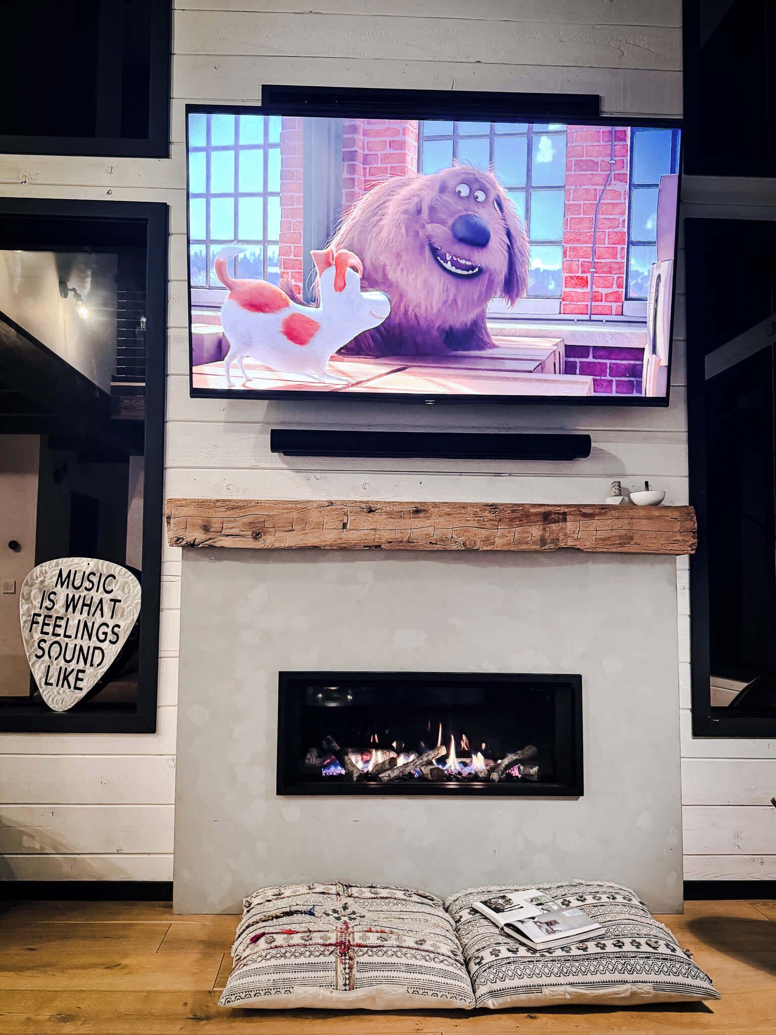 A living room with a flat-screen TV mounted above a fireplace, a heart-shaped sign on the wall, and patterned cushions on the floor.