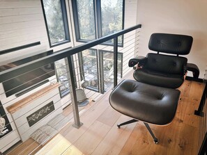 A modern room with a black leather chair, a glass railing, and a view of the outdoors.