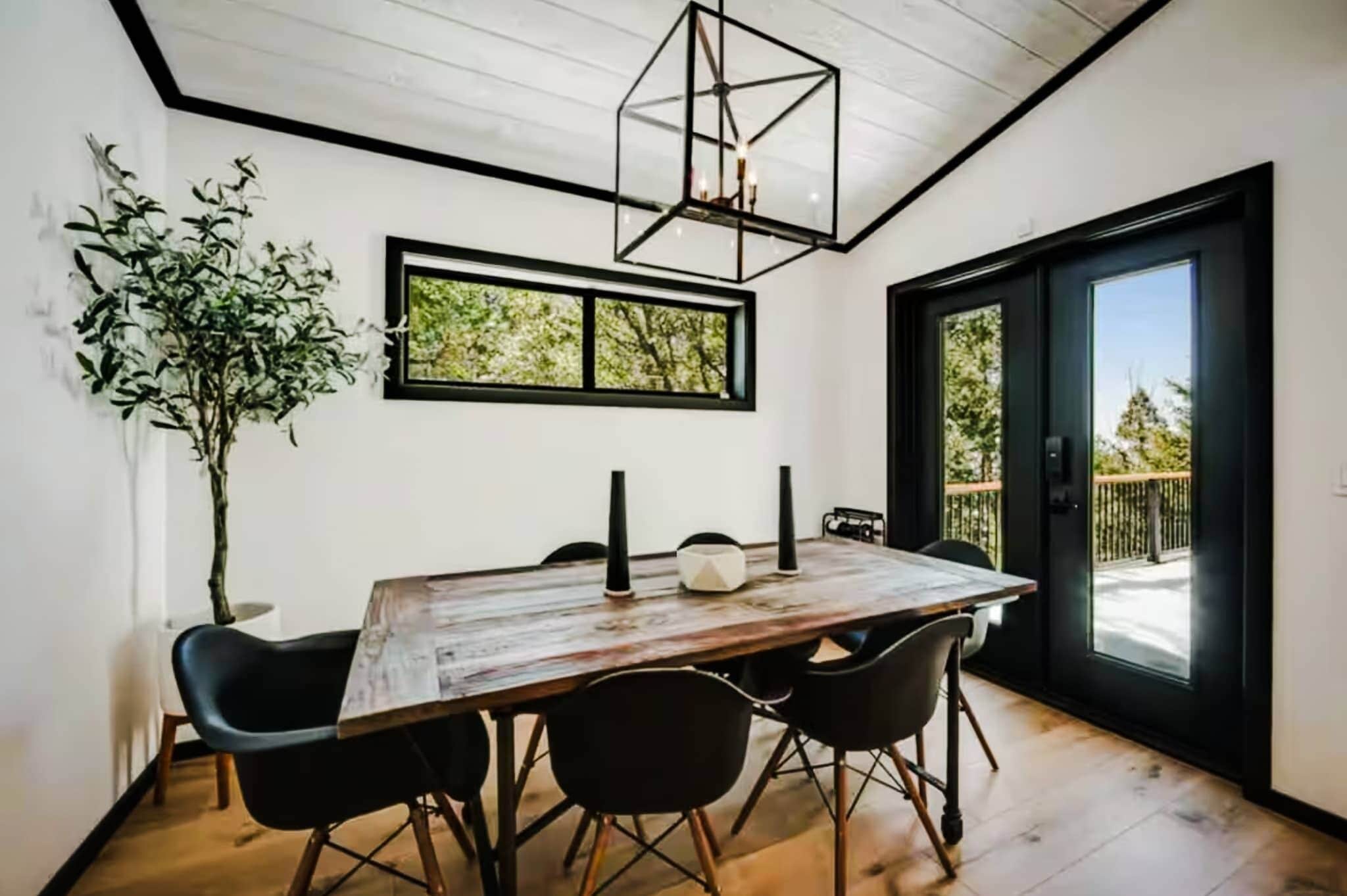 Un coin repas avec une table en bois, des chaises noires, une plante en pot et une vue par la fenêtre.