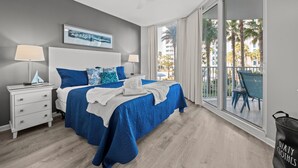 A bedroom with a large bed, a ceiling fan, a nightstand, a window with a view of palm trees, and a balcony with chairs. - 1207 Amazing Lagoon Pool Views ~ Onsite Bar ~ Gym ~ Hot Tub ~ Book Sept 7th (Destin)