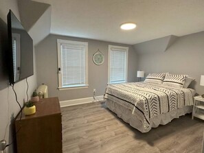 Une chambre à coucher comprenant un lit, une commode, un téléviseur et deux fenêtres équipées de stores.