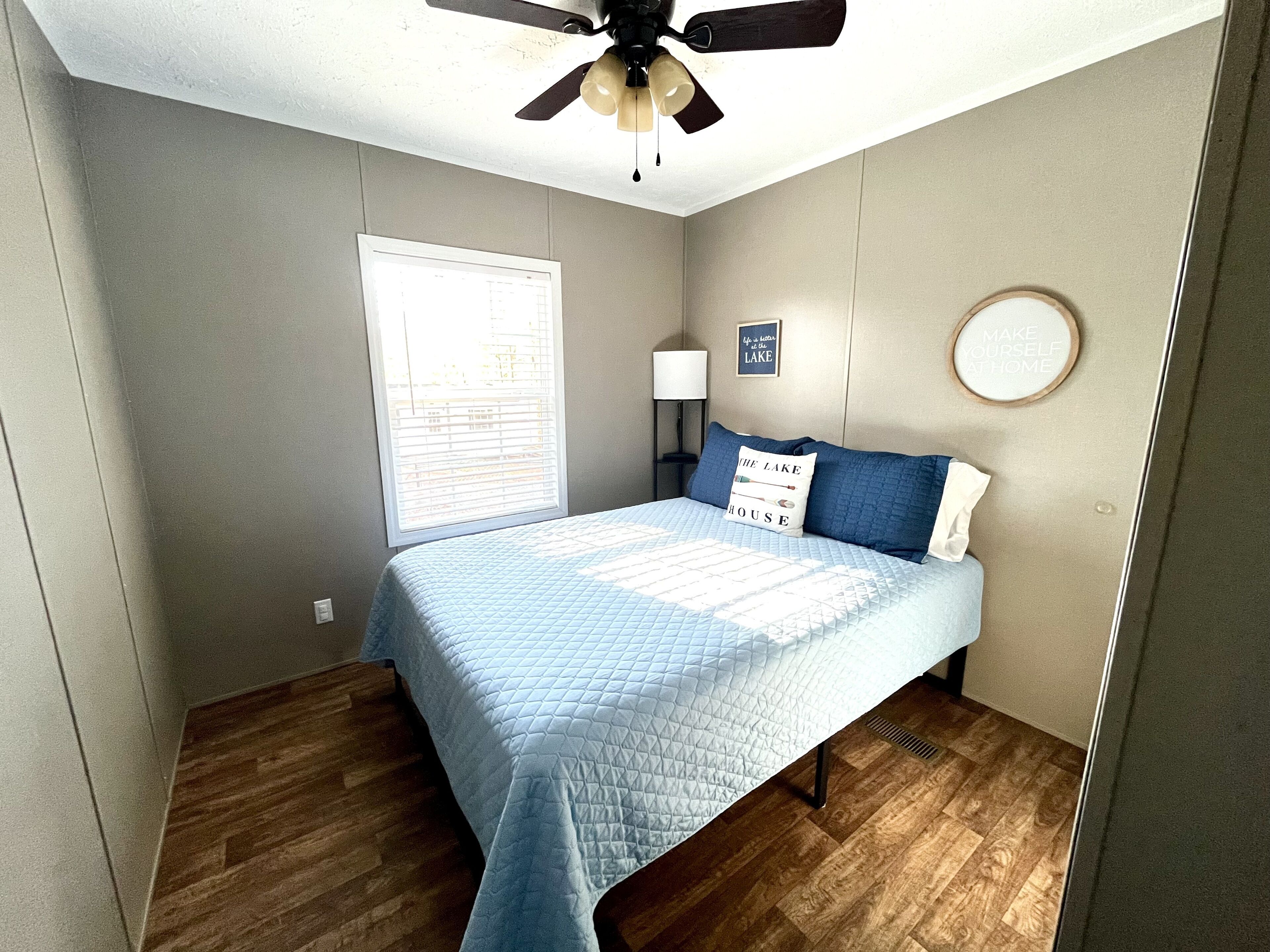 A bedroom with a bed, a ceiling fan, a window, a wall-mounted lamp, and a motivational quote on the wall.