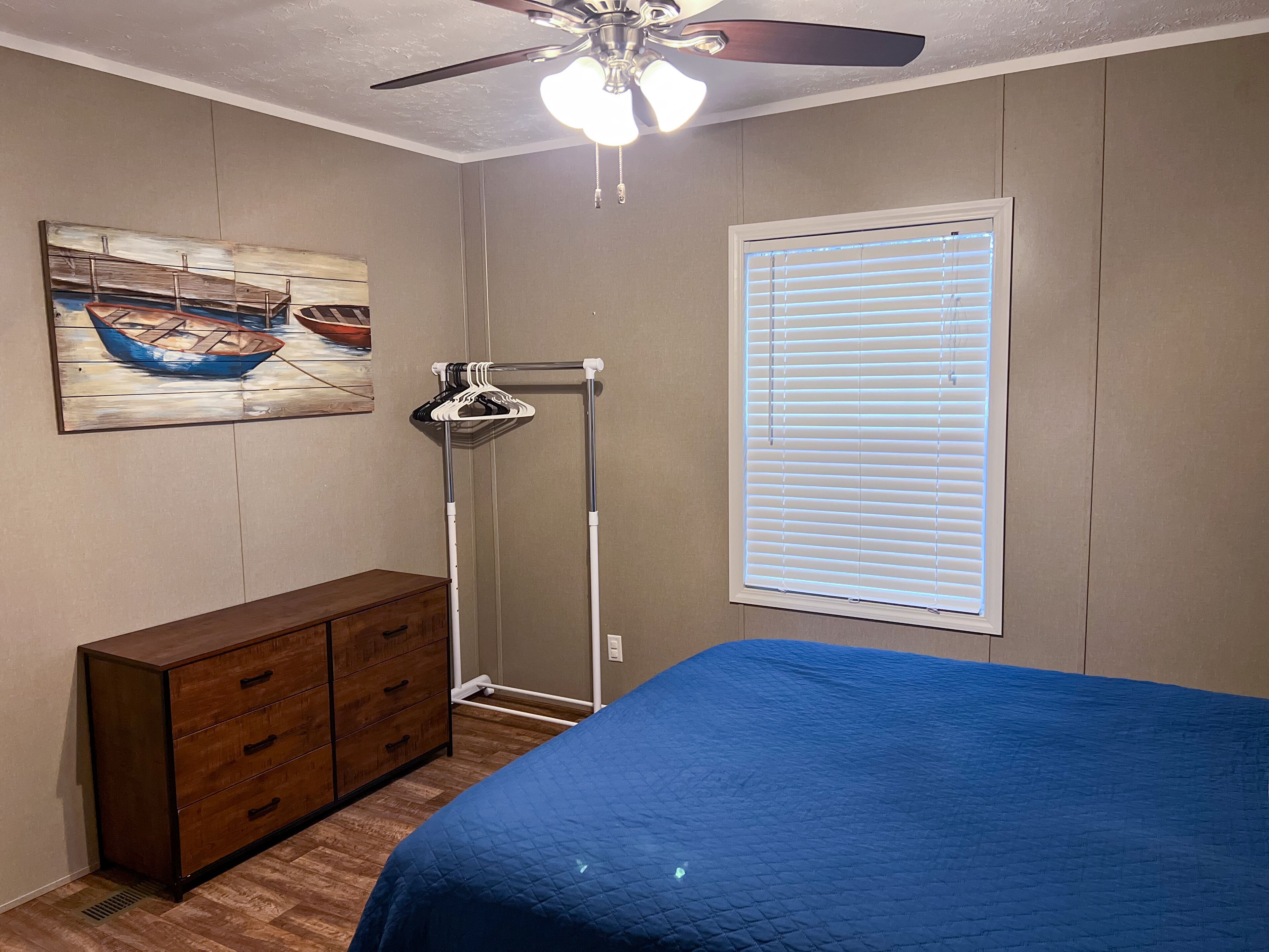 A bedroom with a bed, dresser, ceiling fan, window with blinds, and a painting of a boat.