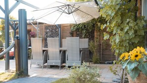An outdoor seating area with wicker chairs and a large umbrella, surrounded by greenery and a stone wall. - Großes Meer (Südbrookmerland)