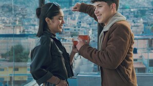 Una pareja de pie junto a una ventana con vistas a un paisaje urbano y montañas al fondo.