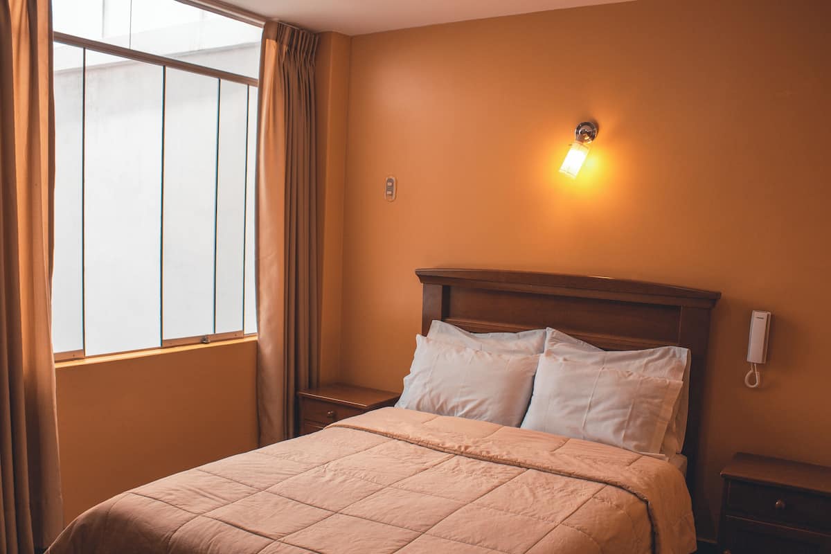 A hotel room with a bed, a nightstand, a wall-mounted phone, and a window with curtains.