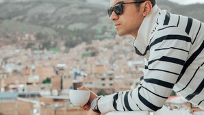 Una persona apoyada en la barandilla de un balcón, sosteniendo una taza, con vistas a un paisaje urbano con edificios y colinas al fondo.