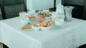 Una mesa de desayuno servida con tazas y platillos blancos, una cafetera, una jarrón de leche y un bol de pasteles.