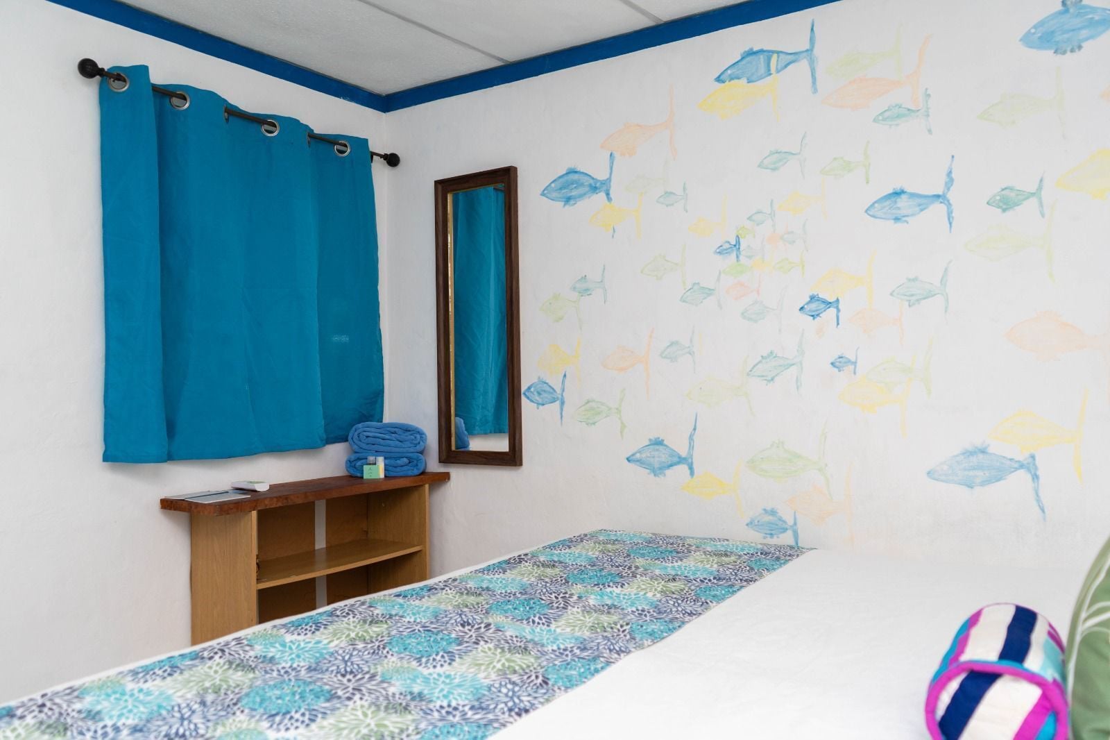 Una habitación con cama, una cortina azul, un espejo, una estantería con objetos y una pared decorada con calcomanías de peces.
